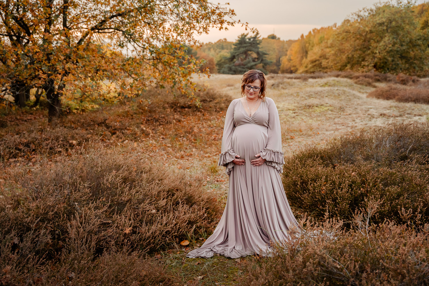 Frau mit Babybauch beiihrem Schwangerschaftsshooting in den Boberger Dünen bei Sonnenuntergang im Herbst ihren bauch liebevoll haltend in einem Babybauchkleid