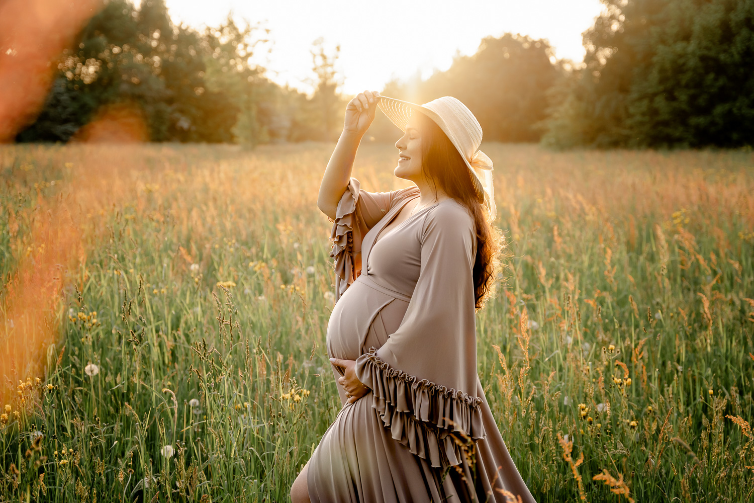 Schwangere Frau bei ihrem Babybauch-Shooting auf einer Wiese bei Sonnenuntergang mit Hut in der Hand und einem Babybauchkleid mit warmer Bildstimmung