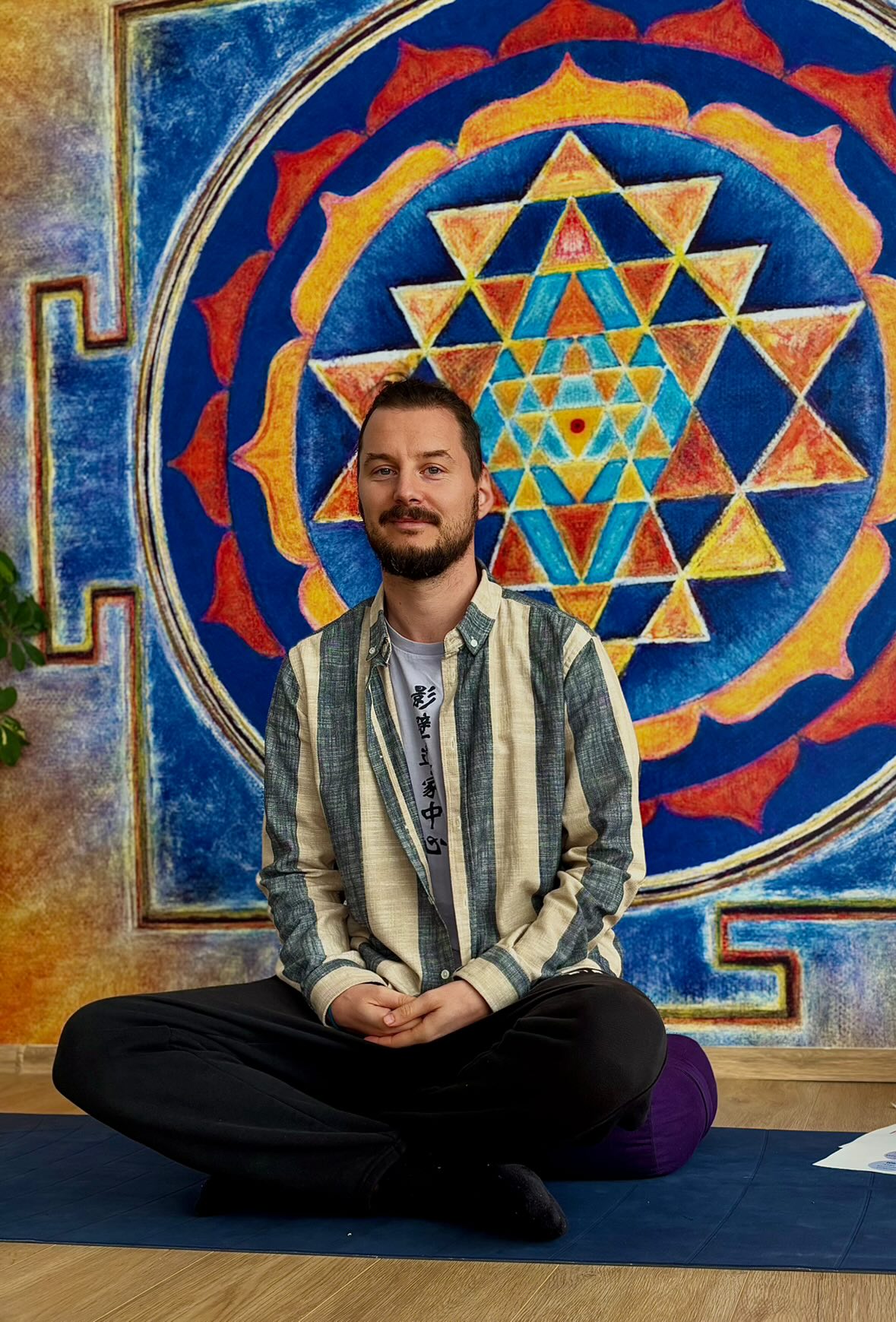 Усмихната, опитна жена-инструктор в началото на 40-те с къса тъмна коса, облечена в мека зелена ленена риза, стои в светло студио с книги и растения, излъчвайки топлота и професионализъм.