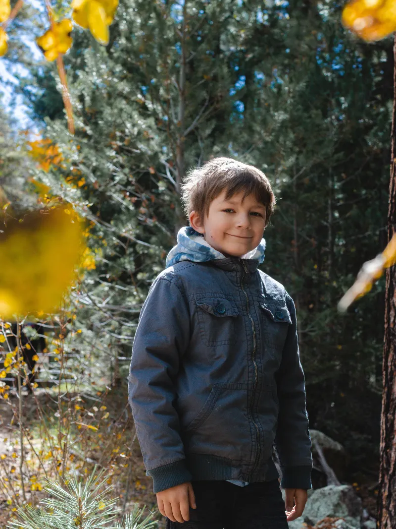 Young boy smiling among evergreen trees during a fall children's portrait session with Andria May Photography in Gilbert, AZ