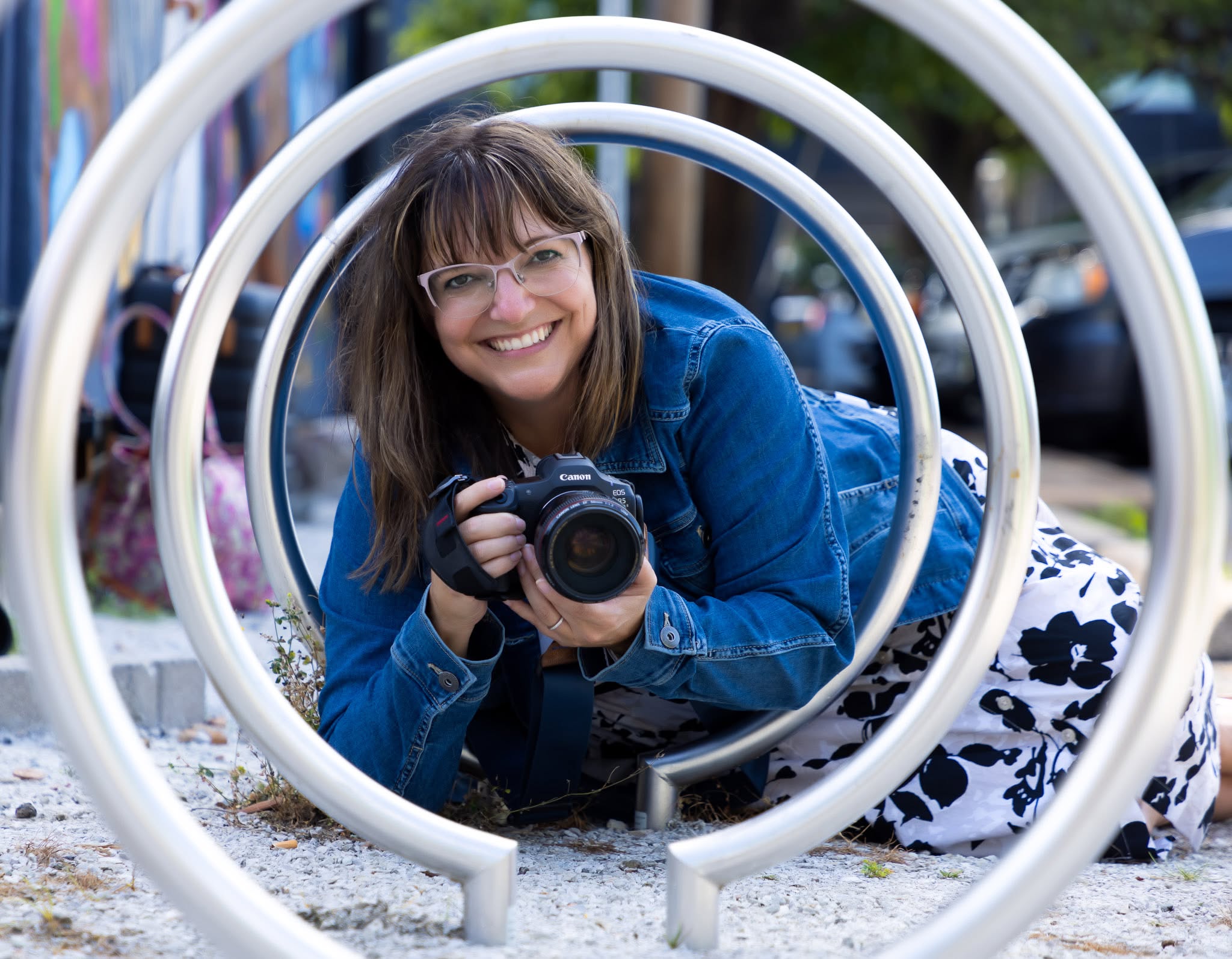Andria May, photographer, crouched behind a Canon camera and shooting through a circular bike rack sculpture in The Loop, St. Louis."