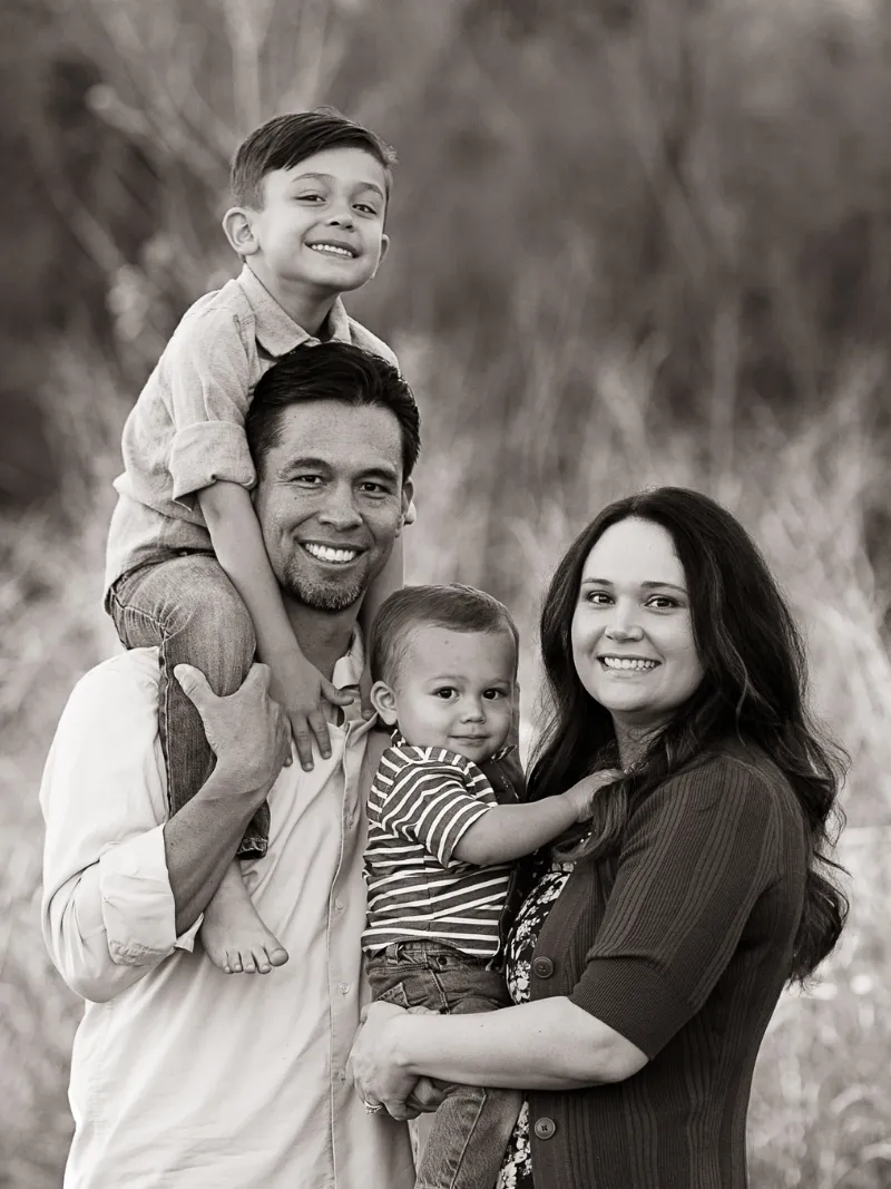 Black and white family portrait of a dad carrying his son on his shoulders while mom holds their toddler outdoors taken by Andria May Photography in Gilbert, Arizona