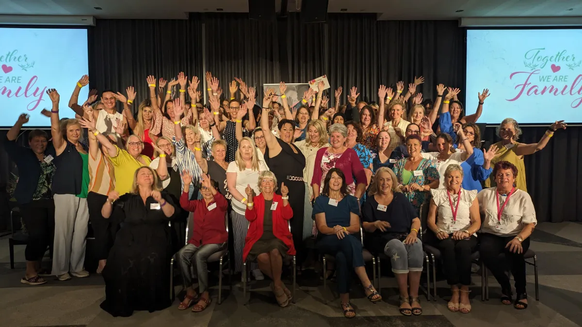 Large group of diverse women smiling in an auditorium, mid-action applause and raised hands, warm natural lighting, candid documentary style emphasizing community energy and hope after transition, photographed from slight elevation.