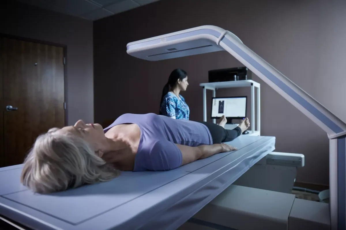 A patient lying on a table, undergoing a DEXA bone density scan. The machine is positioned above the patient, emitting low-dose X-rays.