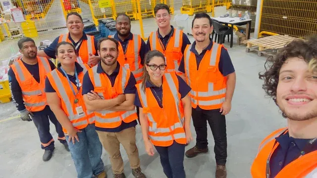 Group of diverse workers in orange high-visibility vests and hard hats, smiling at the camera in a warehouse setting.