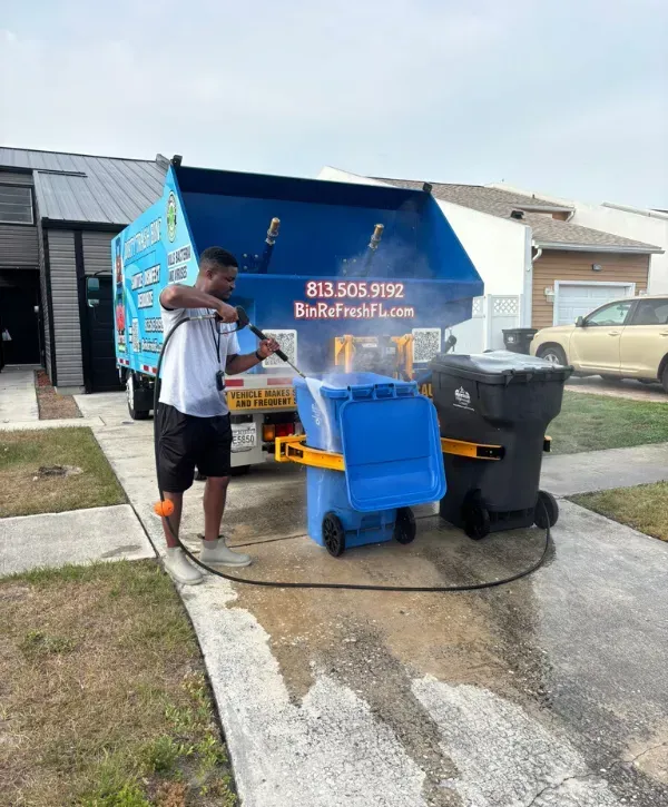 Worker using a pressure washer to clean a blue recycling bin next to a sanitation service truck.