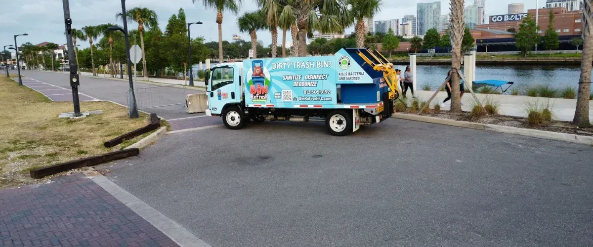 A light blue Bin ReFresh truck with sanitation equipment parked by a canal with a city skyline in the background.
