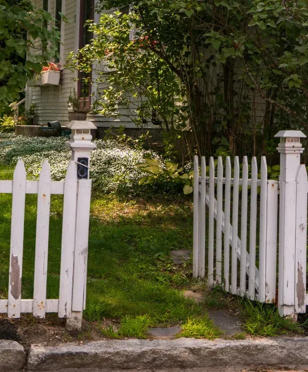 A rustic white picket fence gate stands open in front of a house, revealing a path to the sunlit yard.
