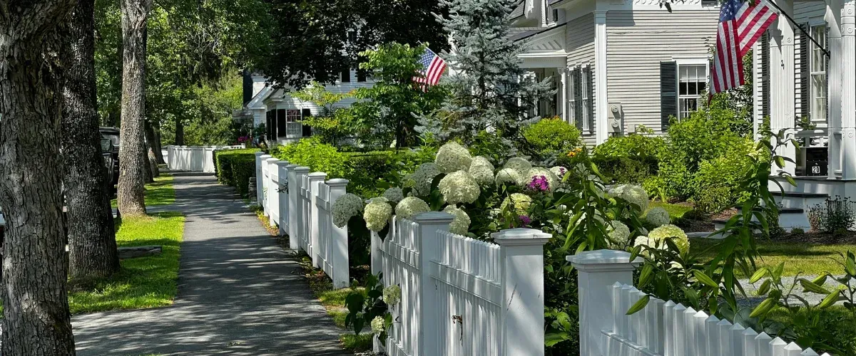 A scenic sidewalk runs alongside a white picket fence adorned with white hydrangeas leading to a New England house.
