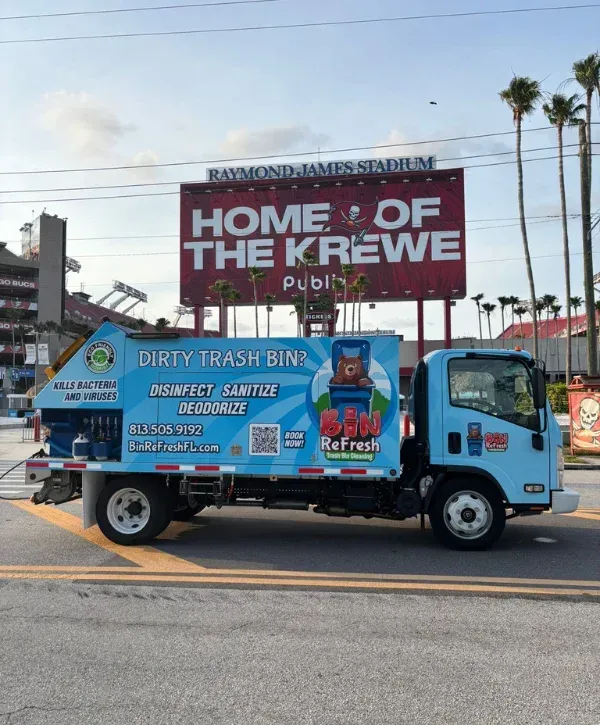 A light blue Bin ReFresh truck parked on a street with Raymond James Stadium and a large billboard in the background.
