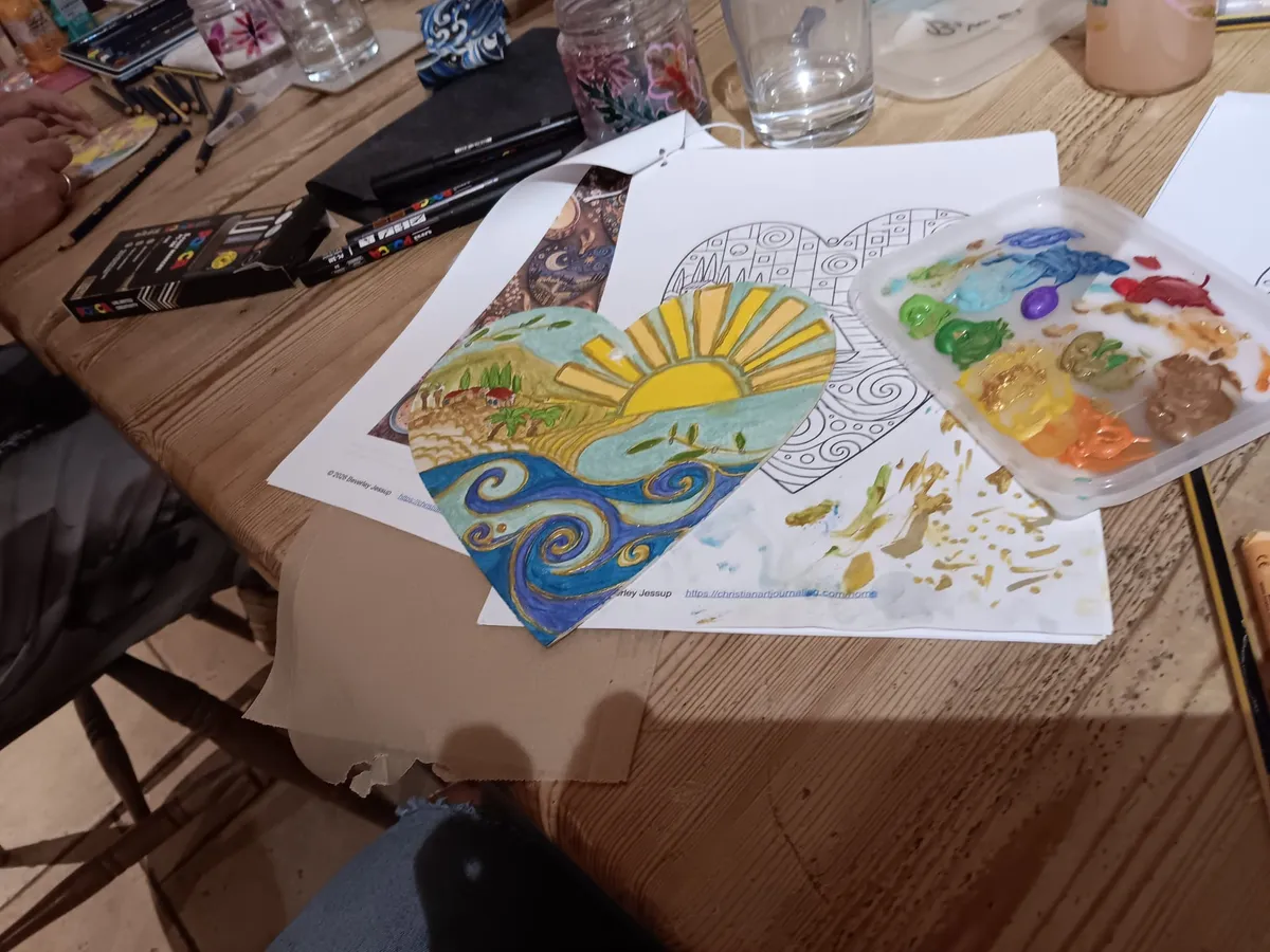 Close-up of hands using a gold paint pen to decorate a heart-shaped illustration on a wooden table, surrounded by art supplies