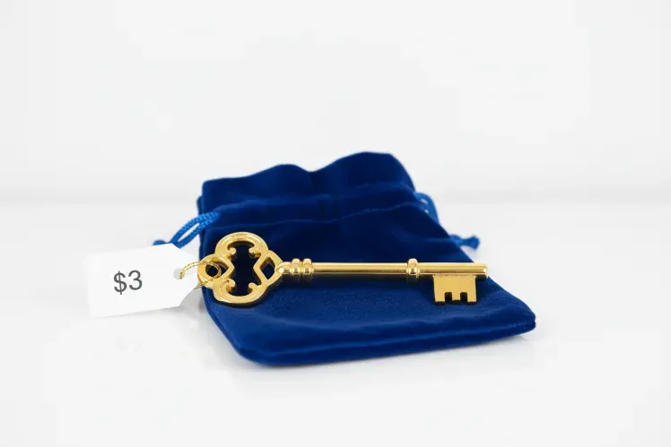 A golden key resting on a royal blue velvet pouch, with a $3 price tag attached. The background is a clean white surface, and the composition is elegant, symbolizing access to wealth and opportunity.