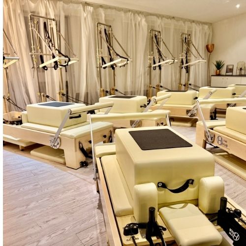 A close-up of a reformer machine in a sunlit studio, with a water bottle and towel placed nearby. The background shows blurred movement of a participant preparing for class, emphasizing readiness and a welcoming, beginner-friendly environment.