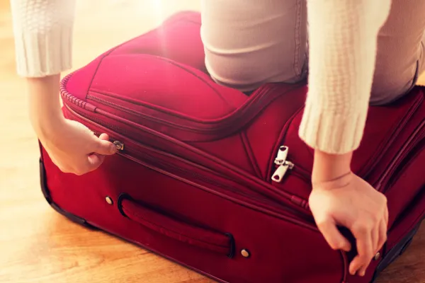 Woman kneeling on an overstuffed red suitcase to force the zipper closed, one hand pulling the zipper pull while the other presses the lid down.