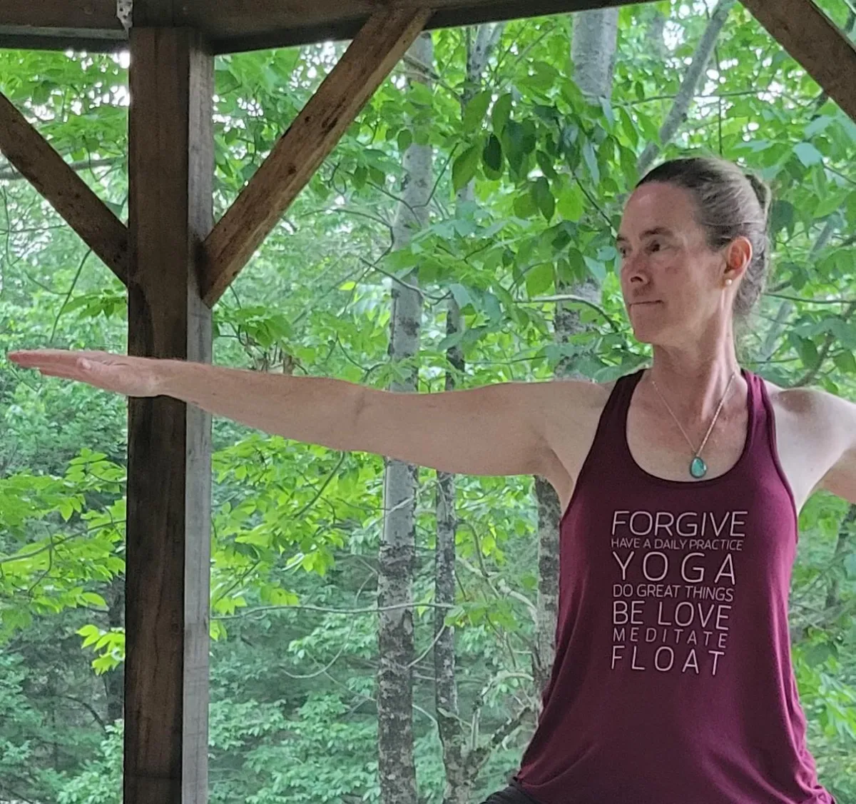 Balance to Bliss Owner Tracy Murdoch performs Warrior 2 pose in a gazebo. There are green trees in the background.
