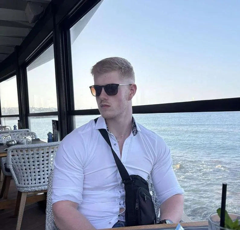 A man with short blond hair and sunglasses sits at a seaside restaurant with large windows, wearing a white button-up shirt and a black crossbody bag. The ocean is visible outside, and a drink with a straw is placed on the table in front of him.