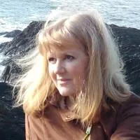 A woman with long, blonde hair is gazing to the side. She is wearing a brown jacket. Behind her, there are rocky formations and sea waves crashing, suggesting a coastal location. The lighting highlights her hair and the serene expression on her face.
