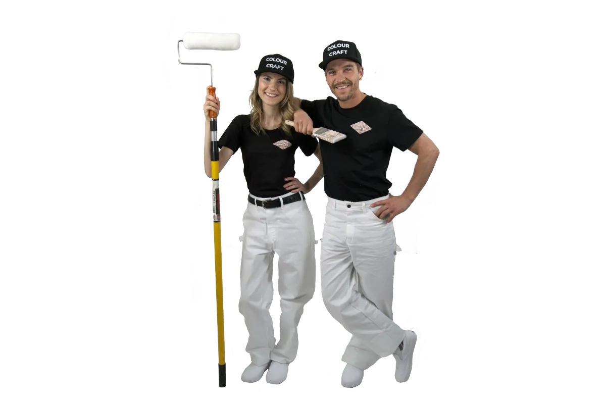 Team of professional painters from Colour Craft, confidently posed with their tools. The woman holds an extended paint roller, while the man sports a brush, both dressed in the company's branded black t-shirts and white pants, ready to deliver high-quality painting services.