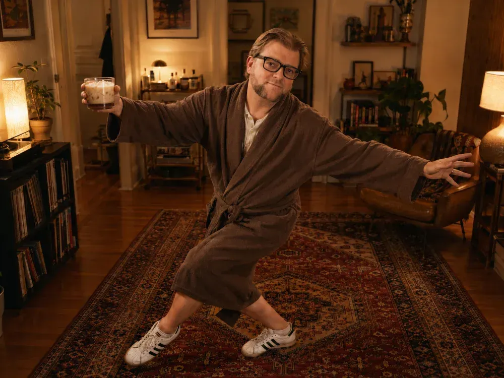 Michael D. Milson doing The Dude's Yoga pose on his rug, wearing his robe, and holding a White Russian