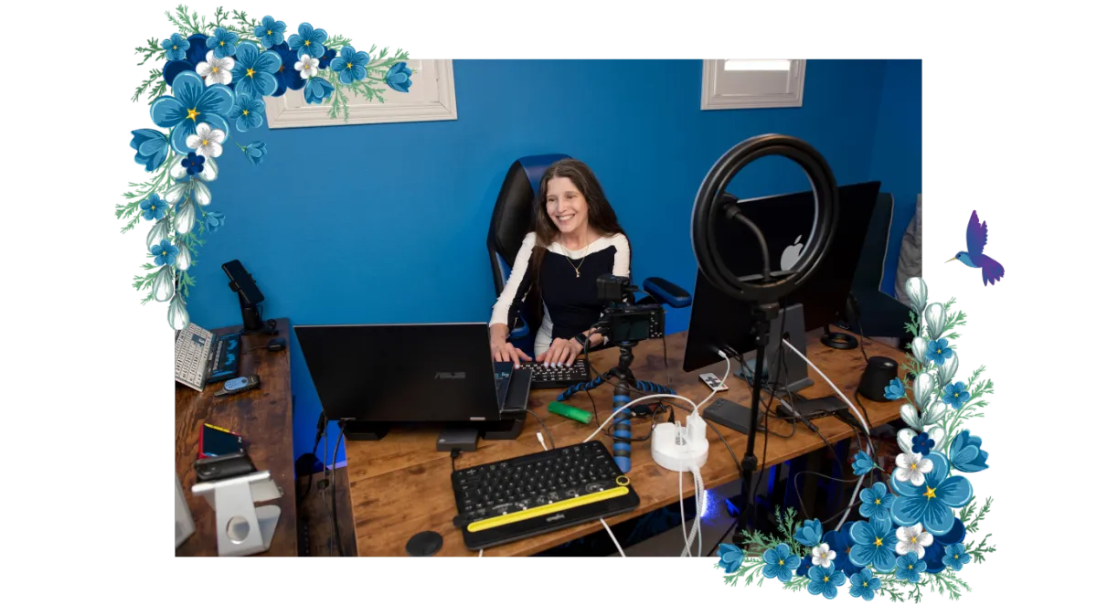 Victoria is smiling while working at a desk in a home office, seated in a blue ergonomic chair. She is wearing a white blouse and using a computer with an accessible keyboard. Bookshelves and technology equipment in the background reflect her role as an assistive technology educator, coach, and advocate.