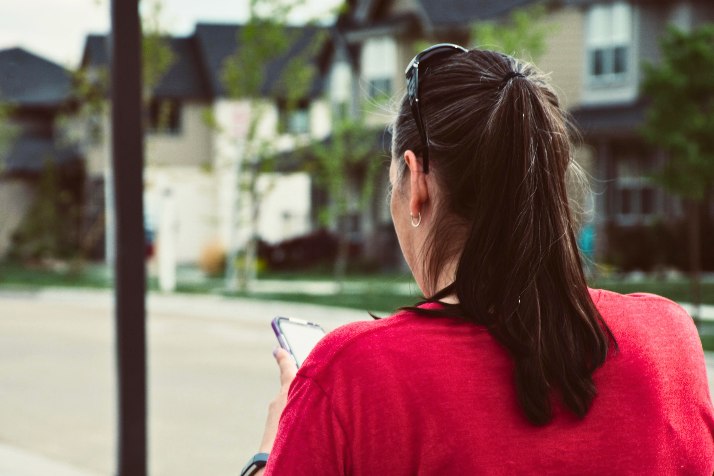 A woman wearing a red shirt looks at her phone in front of a neighborhood sidewalk. There's a row of houses in the background.