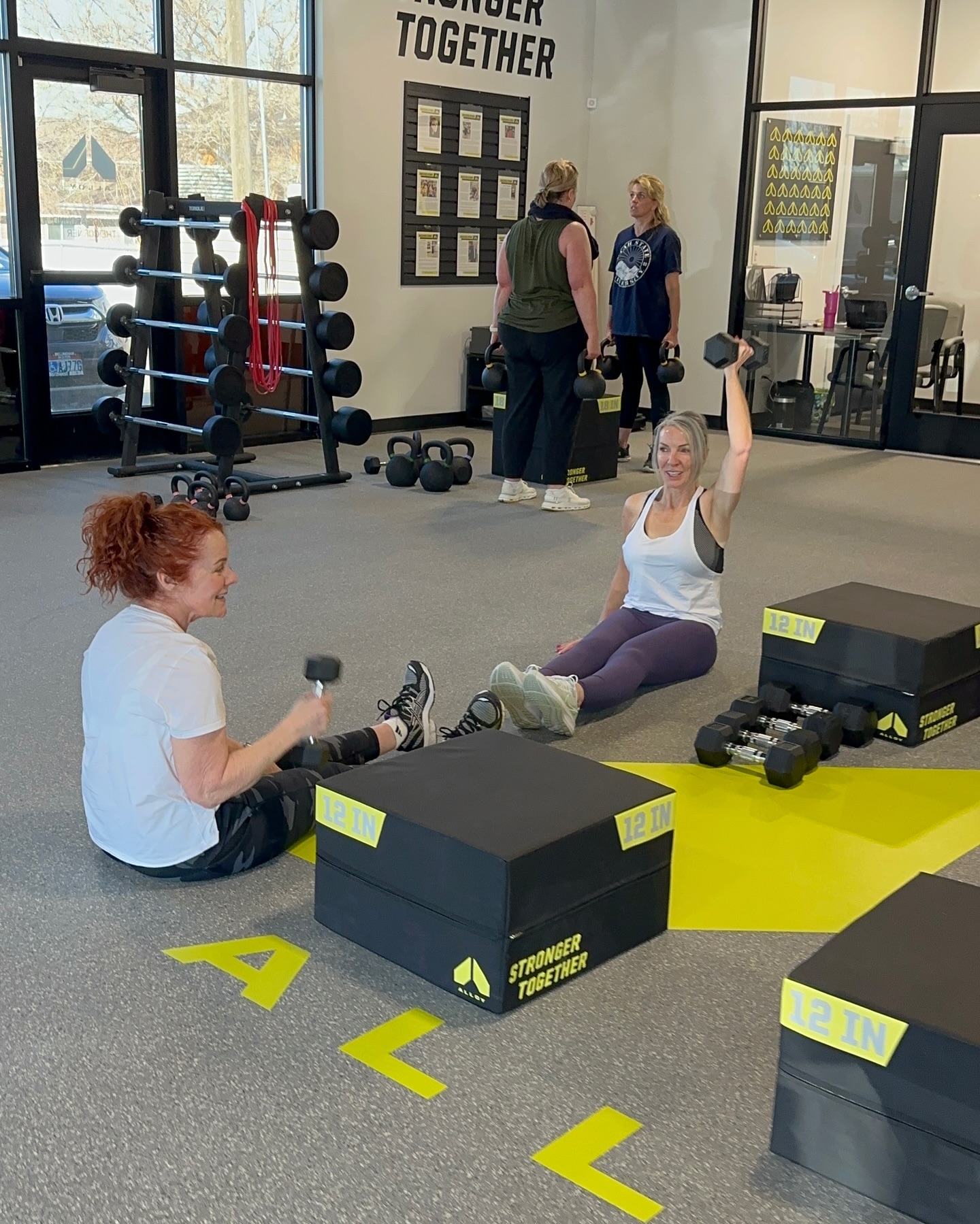  Small group strength training session at Alloy Personal Training Kaysville. Clients working with dumbbells and kettlebells, focusing on strength exercises in a motivating environment.