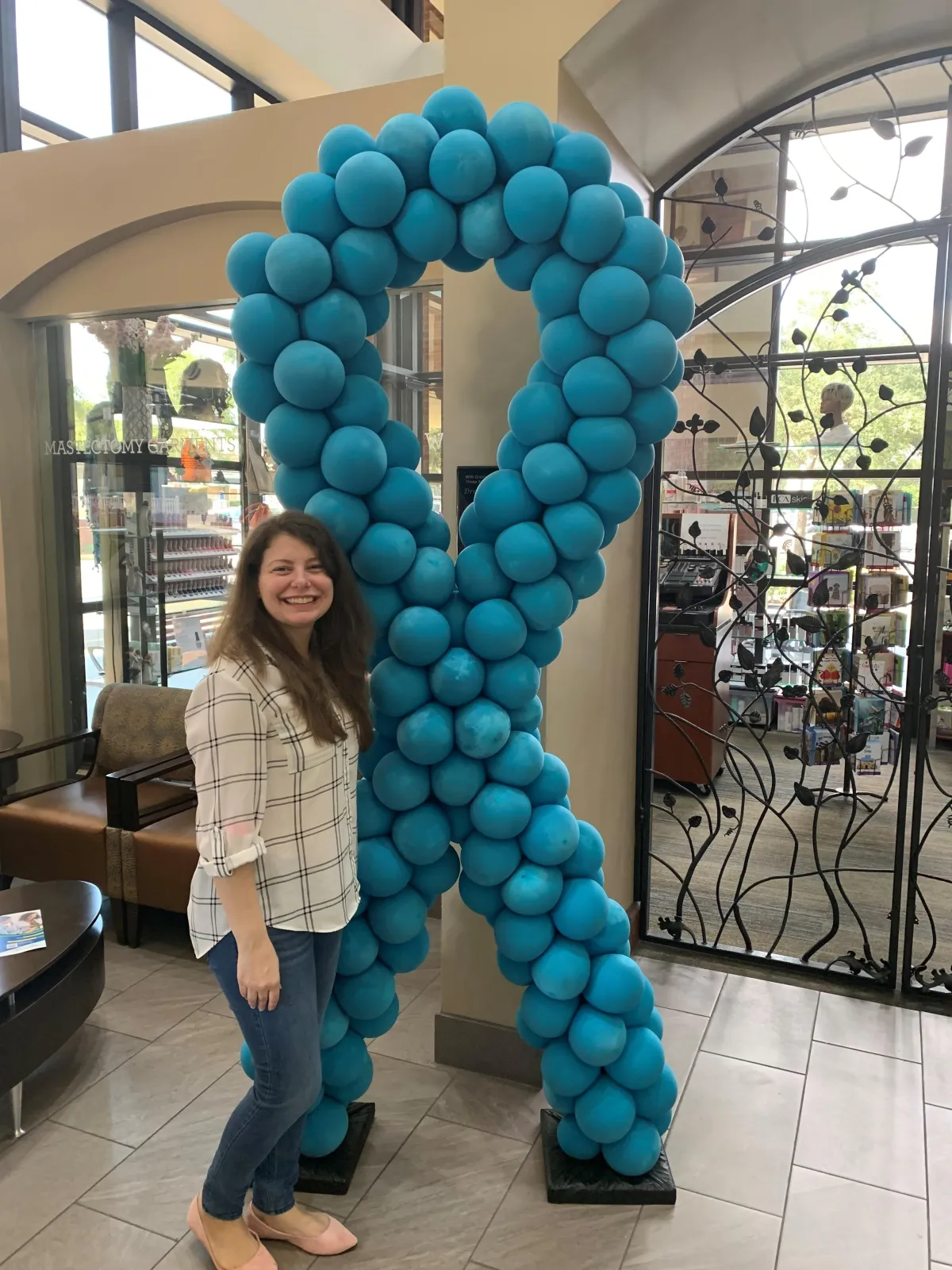 Smiling woman standing next to a large cervical cancer awareness ribbon made from teal balloons, displayed in a medical or public setting to raise awareness for cervical cancer support.