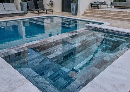 Contemporary backyard pool with integrated spa, featuring sleek gray tile design, lounge seating, and elevated patio steps.