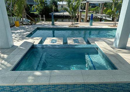 Modern swimming pool with raised spa featuring blue tile accents, overlooking a canal with tropical landscaping and dock access.