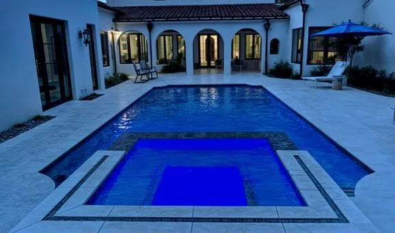Luxury backyard pool with built-in spa, surrounded by white stone decking, lounge chairs, and a Mediterranean-style home with arched windows and terracotta roof.