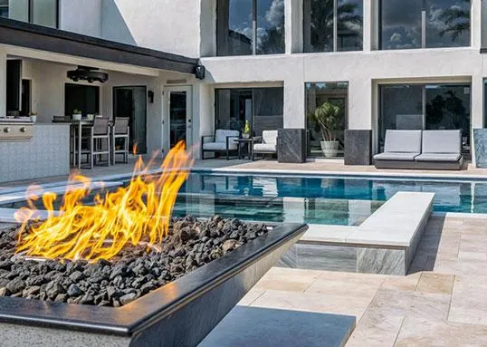 Luxury backyard pool with raised spa, modern lounge seating, and a fire feature in the foreground beside outdoor kitchen and bar area.