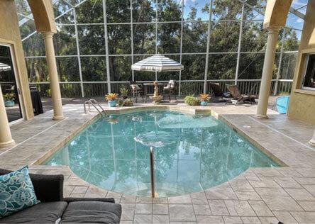 Screen-enclosed backyard pool with rounded edges, surrounded by stone tile deck, lounge chairs, and outdoor dining area under umbrella.