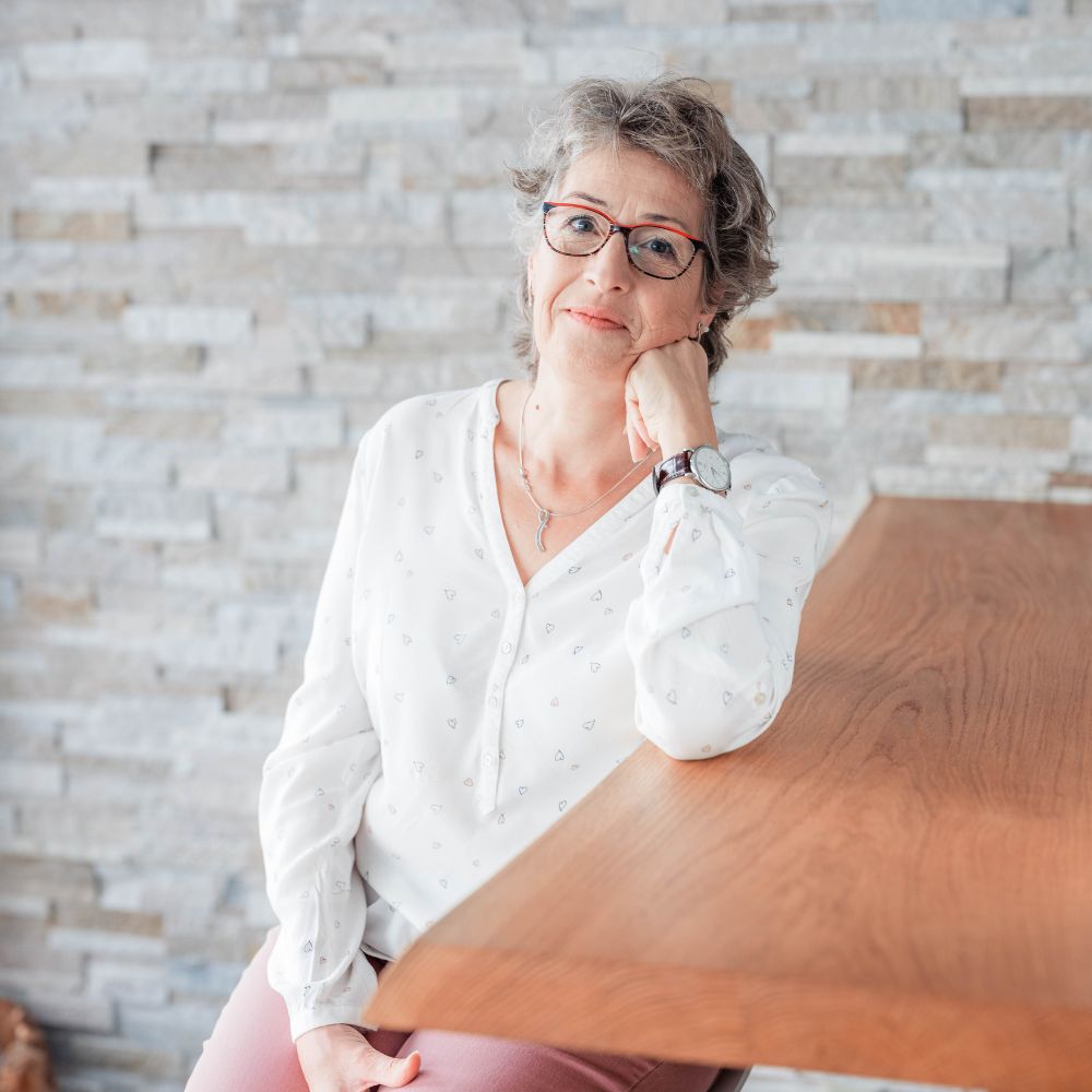 Claudia Hausheer sitzt am Holztisch vor einer hellen Steinwand, Kinn auf der Hand, ruhig und präsent