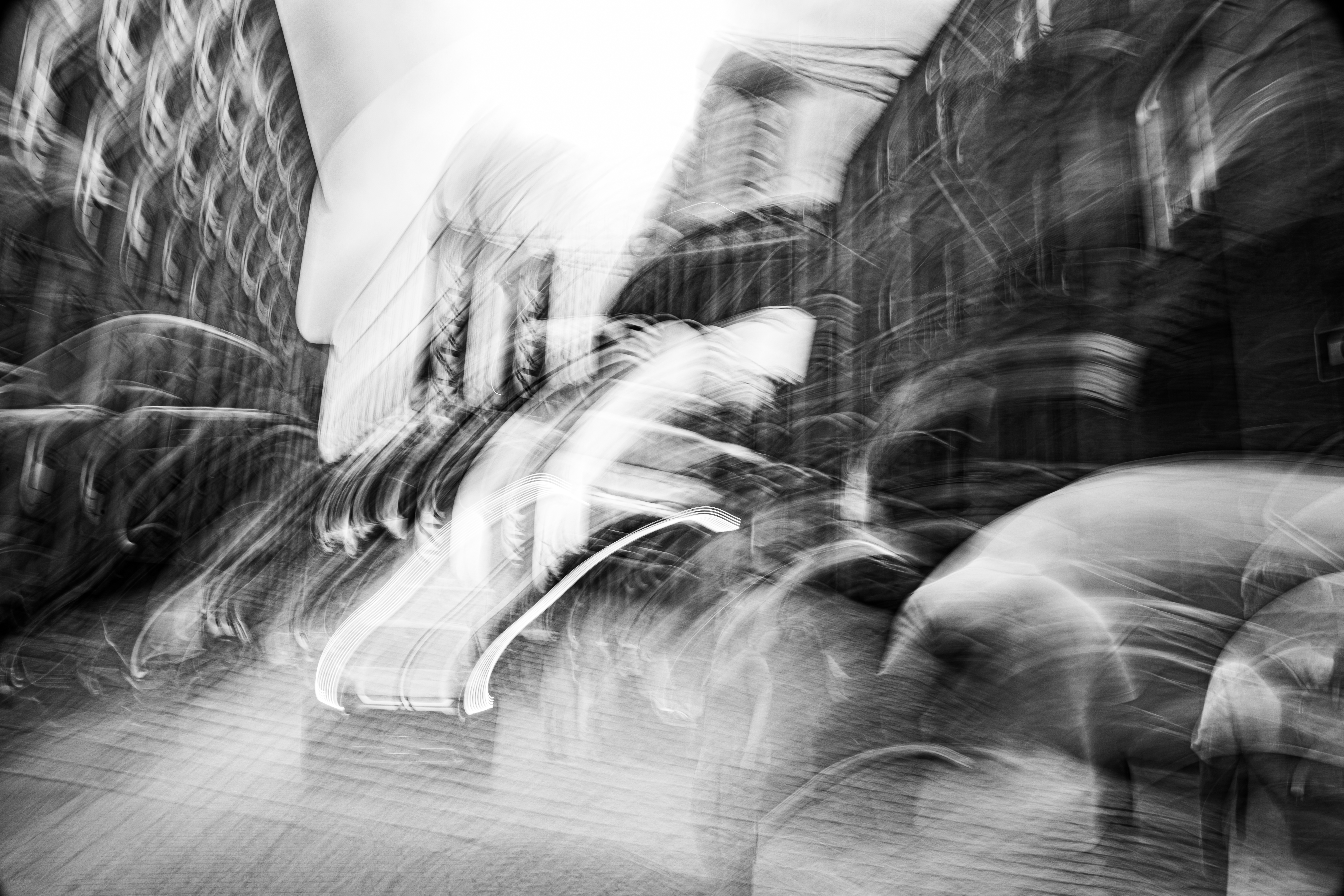 Bold sweeping gestural camera movement through DUMBO Brooklyn in mid-August heat, Brooklyn Bridge visible in background, figures walking ahead and vehicle approaching, buildings pressing in from either side, monochrome ICM fine art photograph by Angie Harker