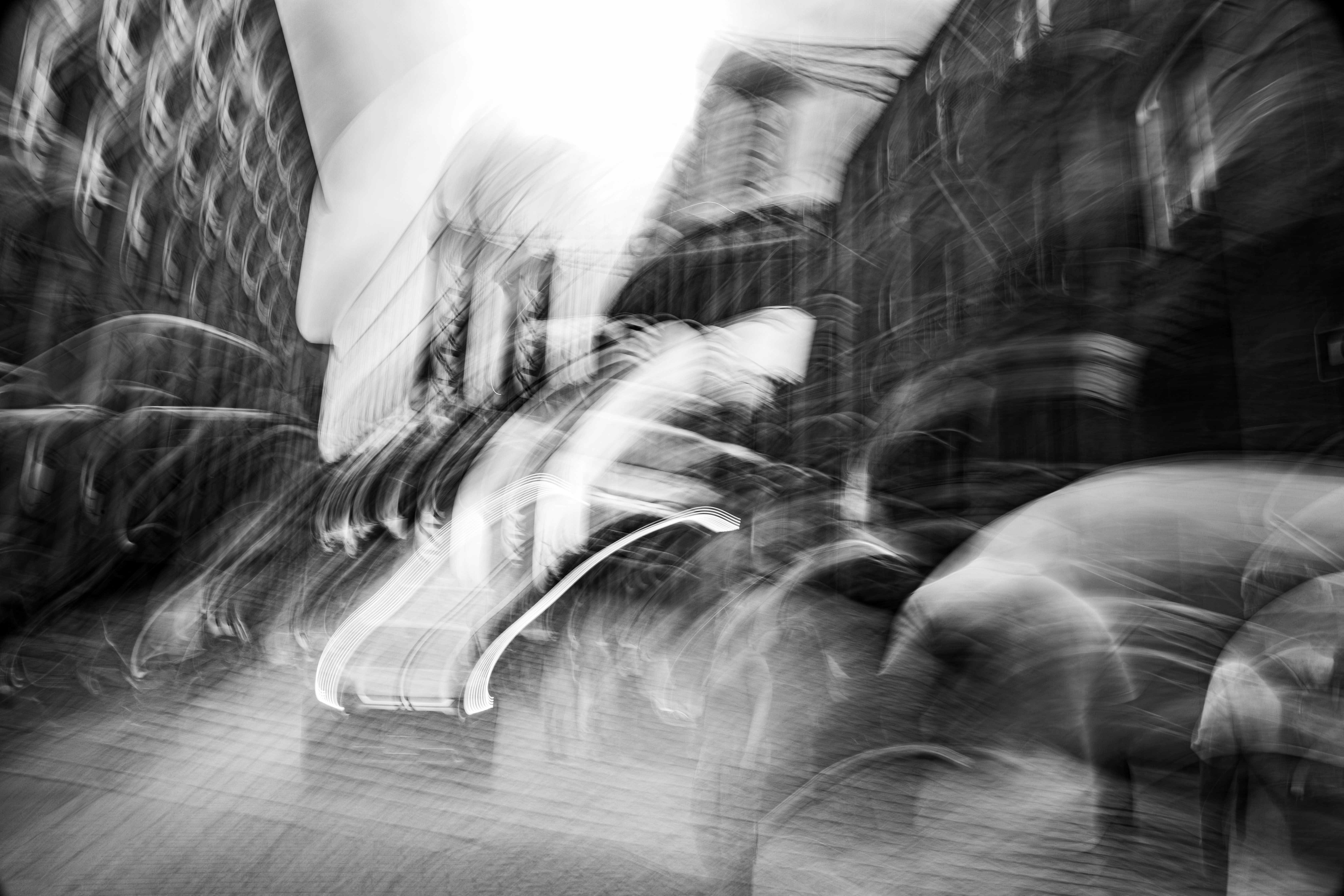Bold sweeping gestural camera movement through DUMBO Brooklyn in mid-August heat, Brooklyn Bridge visible in background, figures walking ahead and vehicle approaching, buildings pressing in from either side, monochrome ICM fine art photograph by Angie Harker