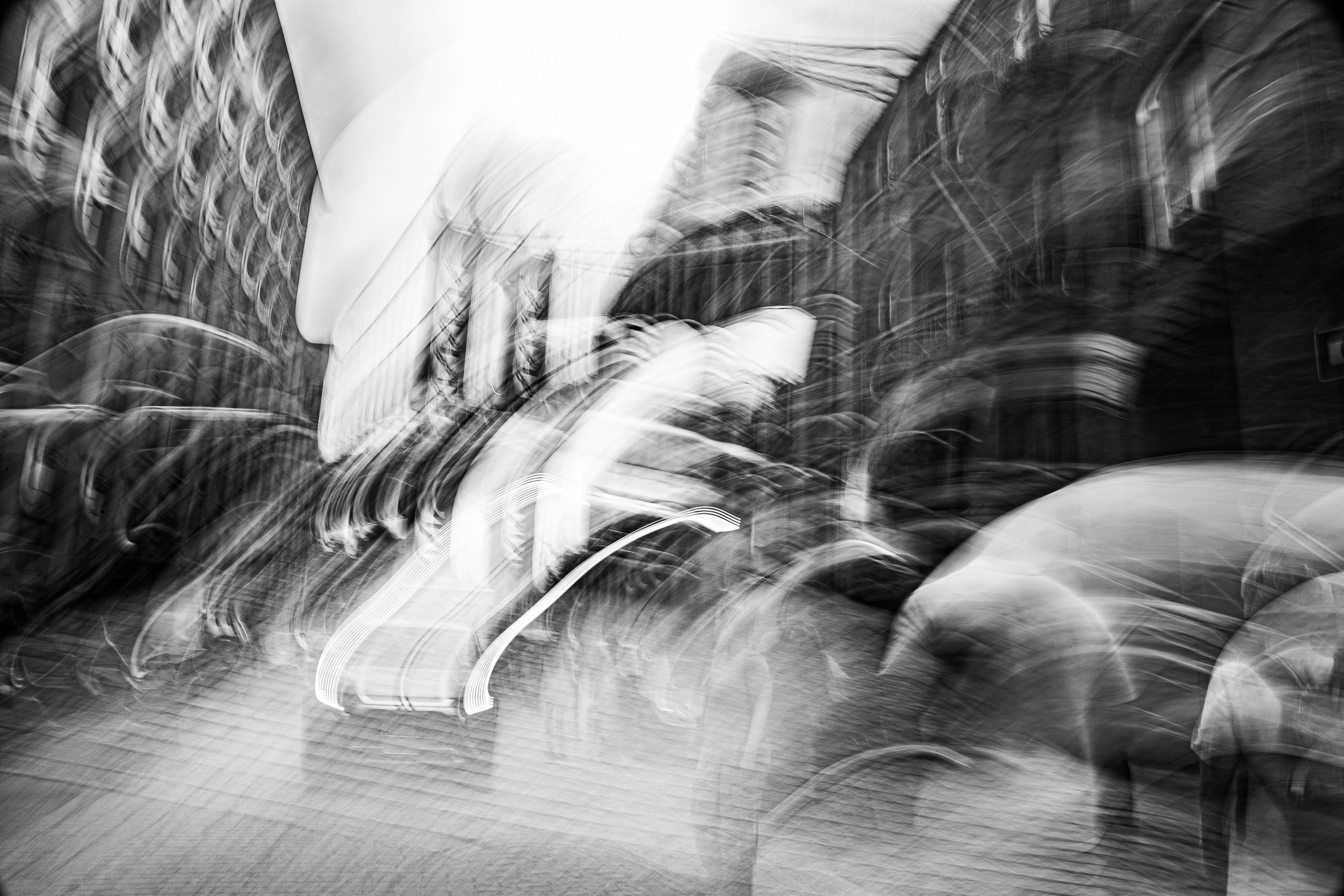 Bold sweeping gestural camera movement through DUMBO Brooklyn in mid-August heat, Brooklyn Bridge visible in background, figures walking ahead and vehicle approaching, buildings pressing in from either side, monochrome ICM fine art photograph by Angie Harker