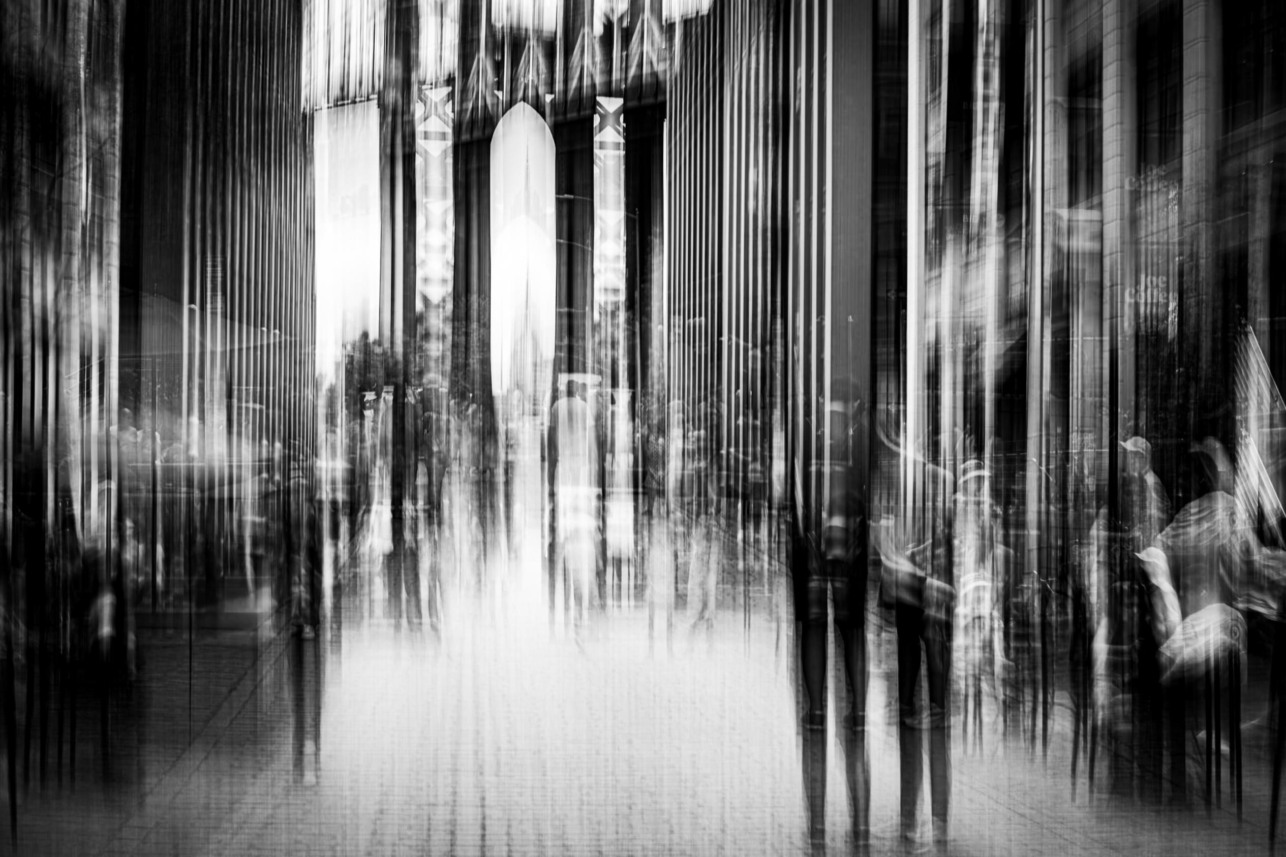 Vertical striations pulling the entire frame upward through DUMBO Brooklyn, figures ghosted within columns of light and dark, Brooklyn Bridge tower visible in the centre background, monochrome ICM fine art photograph by Angie Harker