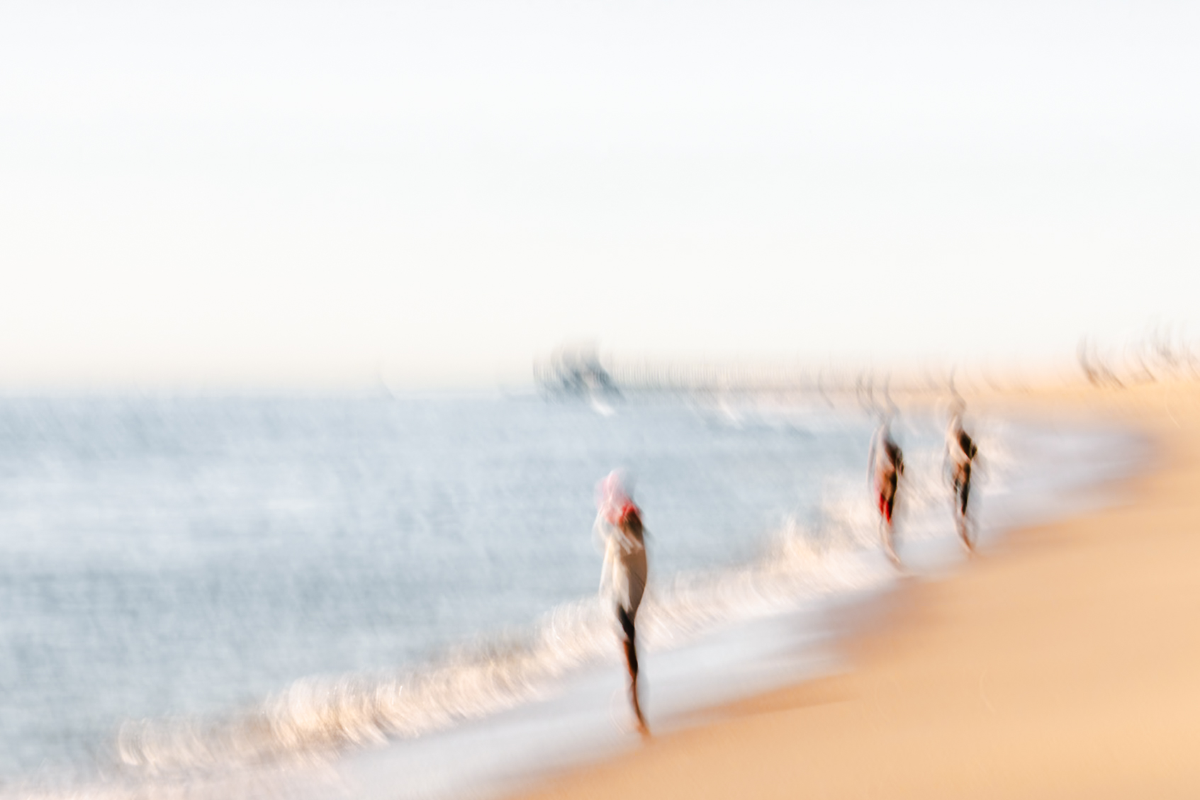 A lone woman standing at the Newport Beach surf's edge, hat in hand, two figures approaching in the distance behind her, warm high key palette, ICM fine art photograph by Angie Harker
