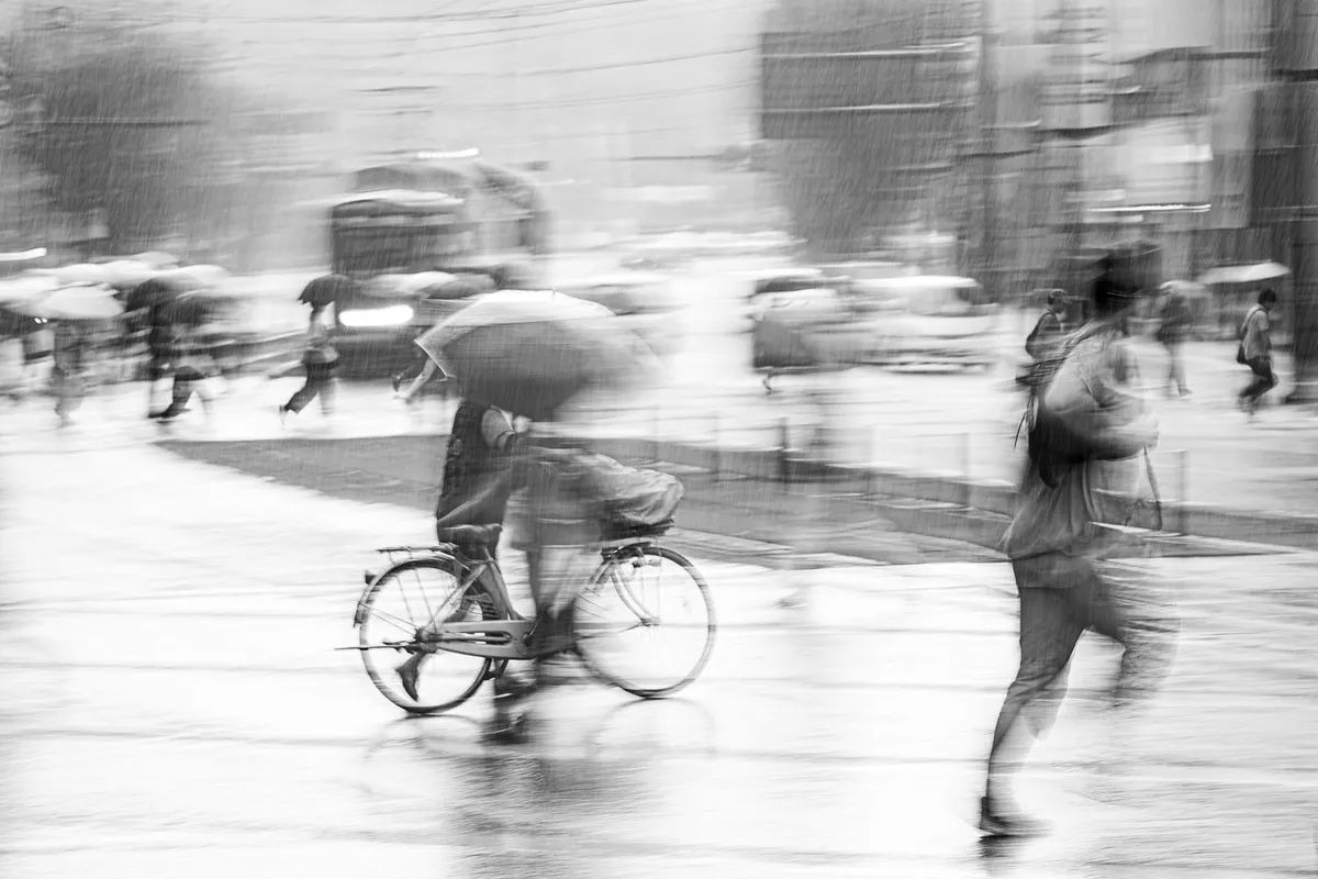A man pushing his bicycle and another sprinting across a Kumamoto street in torrential rain, a layered crowd crossing behind them in opposing directions, monochrome ICM fine art photograph by Angie Harker