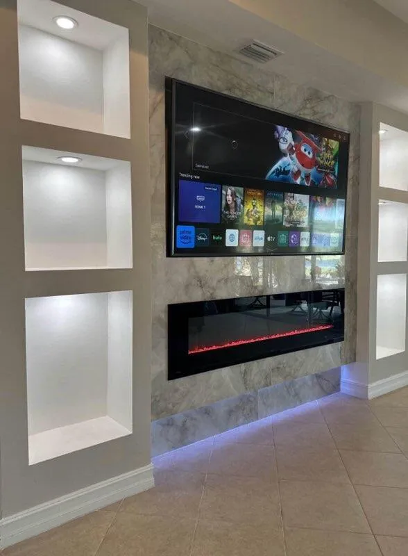 Modern custom media wall with built-in electric fireplace, mounted flat-screen TV, LED-lit stone accent niches, and surround sound speakers in a contemporary living room.