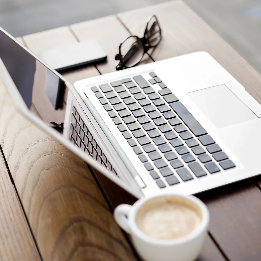 Laptop, coffee mug, and reading glasses on a wooden table.