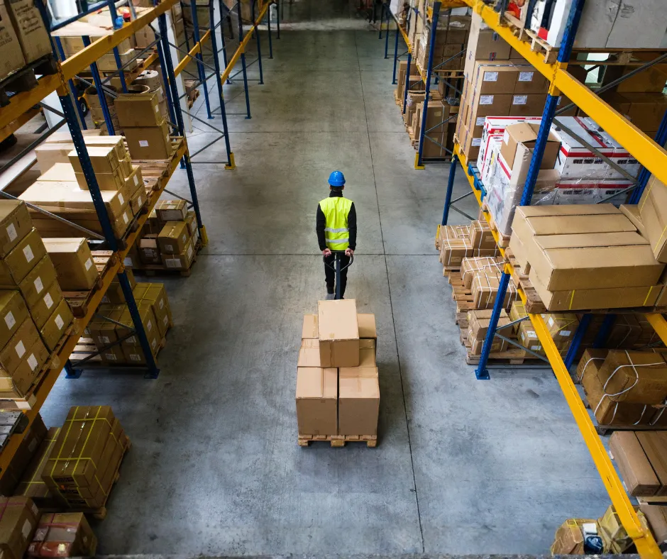 Busy distribution warehouse with stacked pallets, workers using tablets, forklifts, and high shelving.