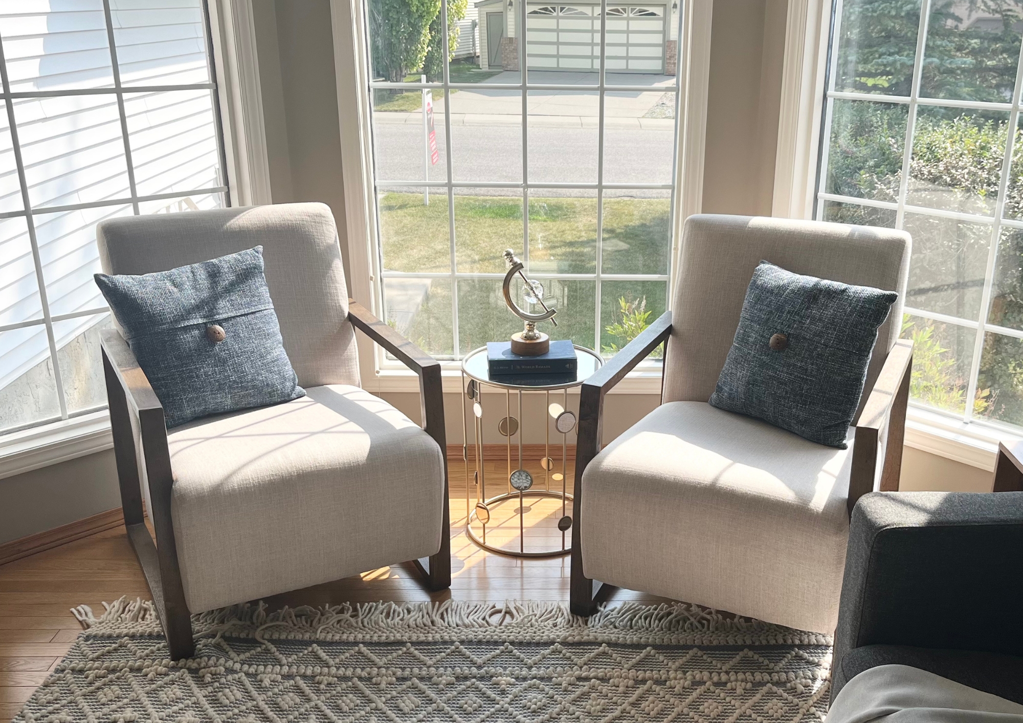 Bright staged seating area with two upholstered chairs, blue accent pillows, and a small round table between them in front of a sunlit window.