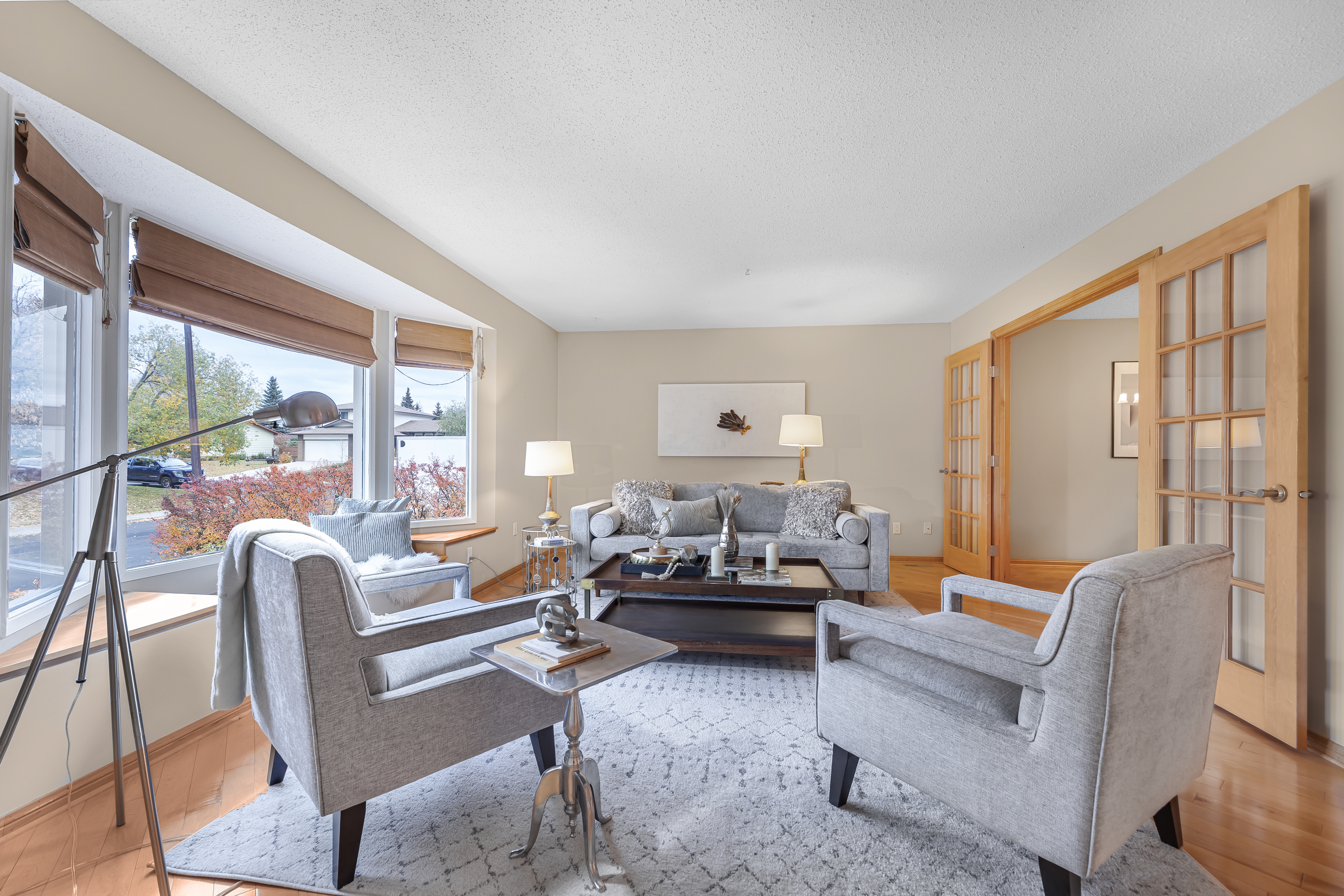 Staged living room with neutral armchairs and sofa around a coffee table, large windows with outdoor views, and soft natural light.