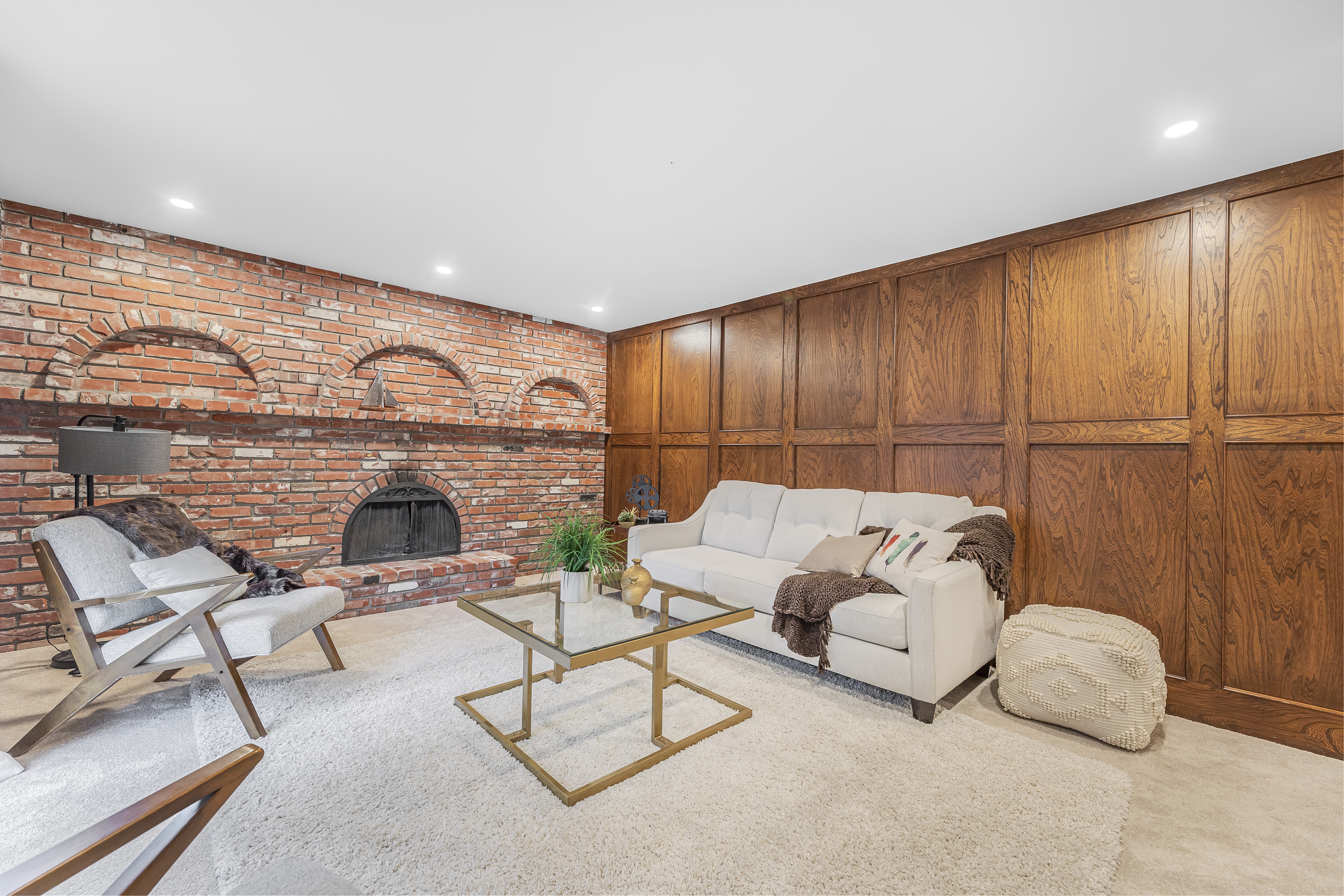 Staged living area with a white sofa, glass coffee table, wood-paneled wall, and brick fireplace with decorative arches.