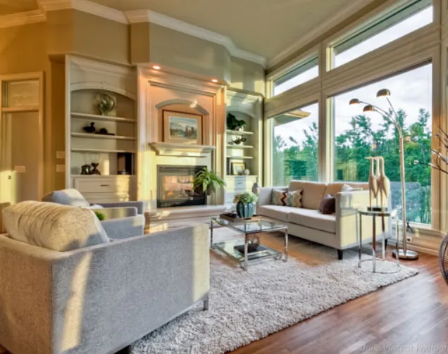 Bright staged living room with a fireplace, built-in shelves, neutral sofas, glass coffee table, plush rug, and large windows bringing in natural light.