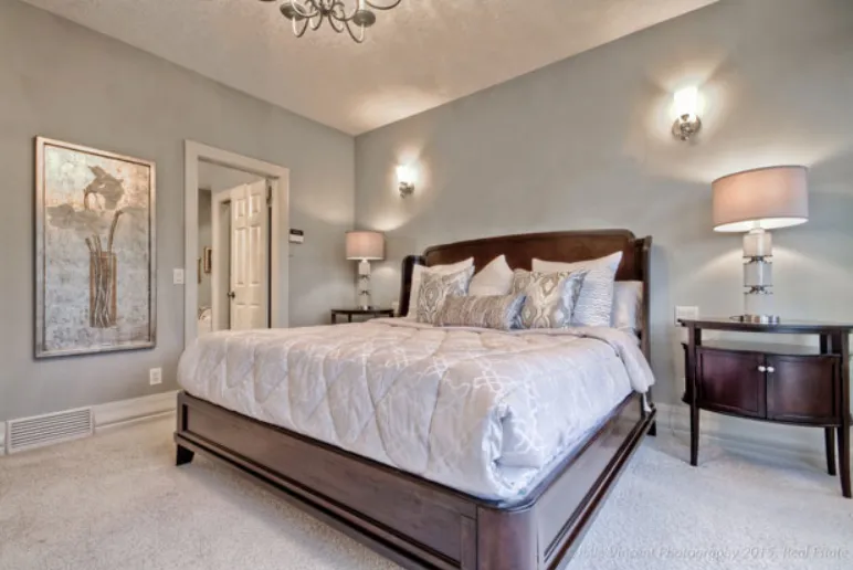 Bright staged bedroom with a wooden bed frame, neutral bedding, decorative pillows, wall sconces, and a bedside lamp.