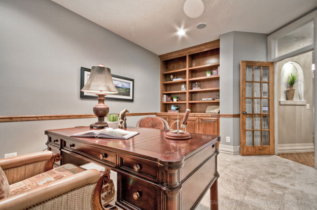 Staged home office with a wooden desk, table lamp, built-in wood shelving, and a glass-paneled door, styled with neutral decor and soft lighting.