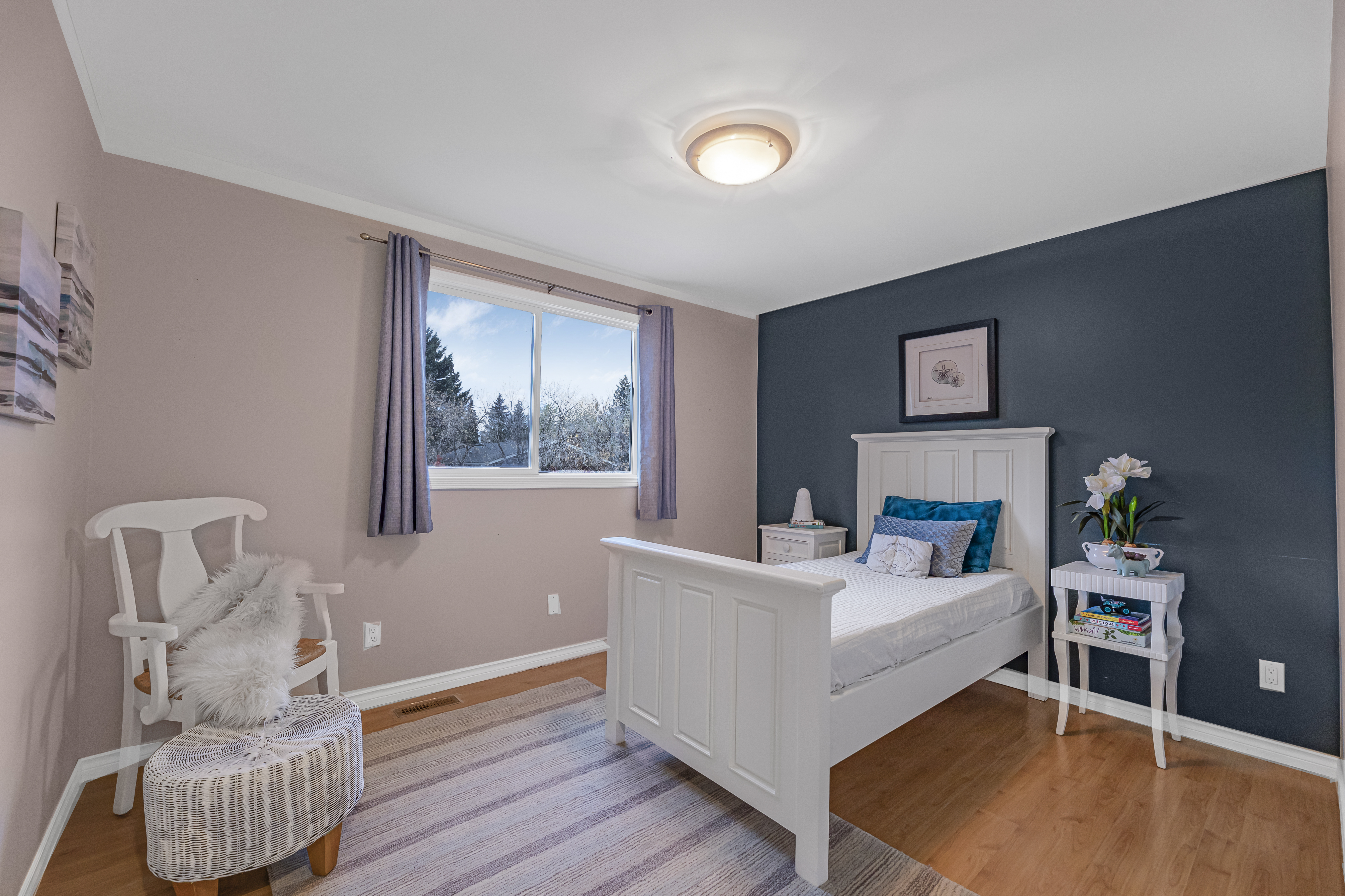 Staged bedroom with a white bed frame, blue accent wall, soft pink walls, and a window bringing in natural light.