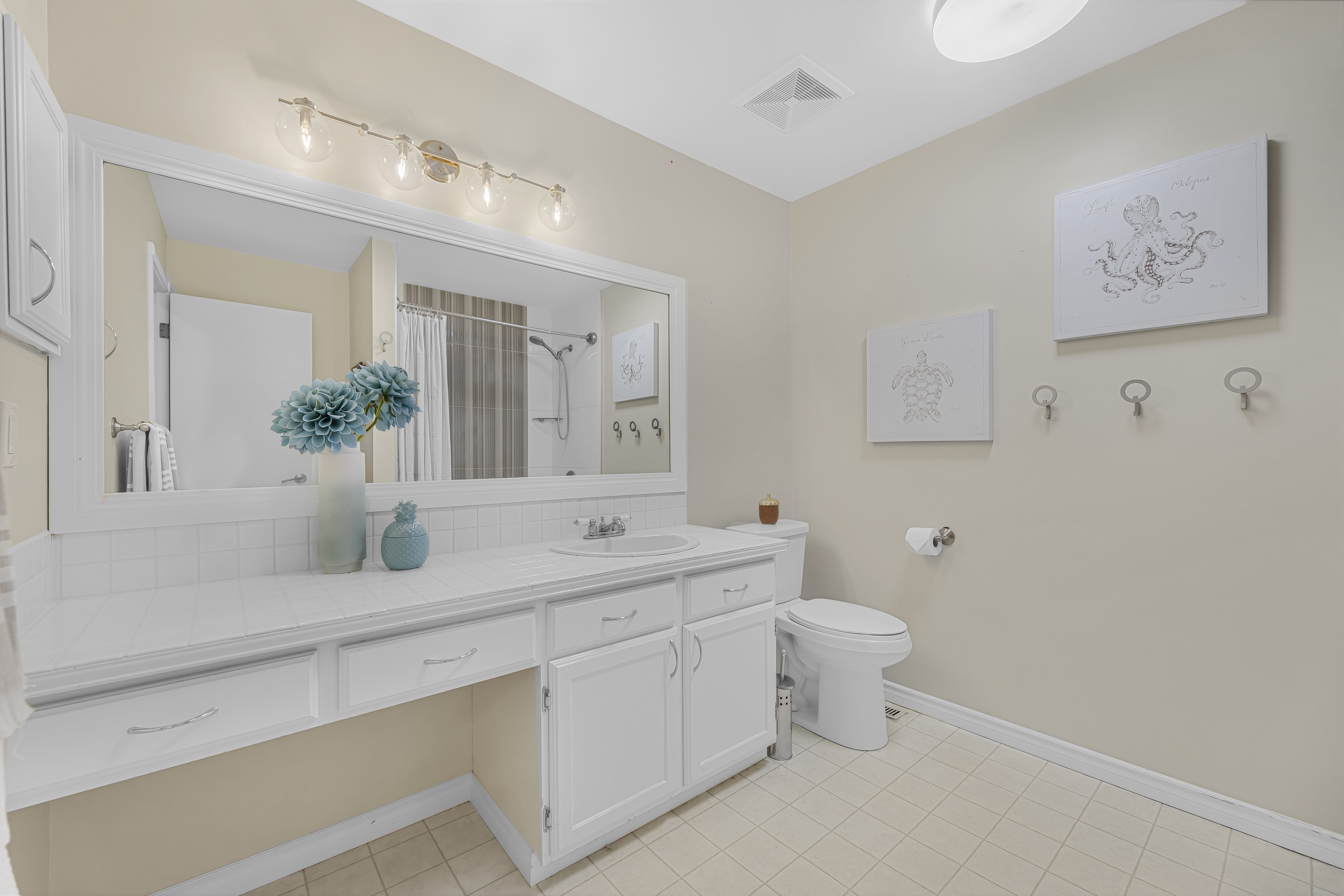 Staged bathroom with a white vanity, large mirror with vanity lights, neutral walls, and simple decor including flowers and wall art.